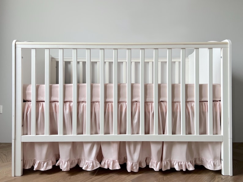 A white wooden crib with a pink mattress cover and ruffled skirt.