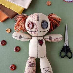 Puede incluir: Una muñeca de trapo hecha a mano con ojos de botón y pelo de hilo. La muñeca tiene una cara, brazos y piernas cosidos, con un parche en forma de corazón en el pecho. Varios suministros de costura, incluyendo botones y retazos de tela, están colocados alrededor de la muñeca.