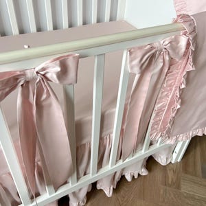 A white crib with pink ruffles and bows on the railings.