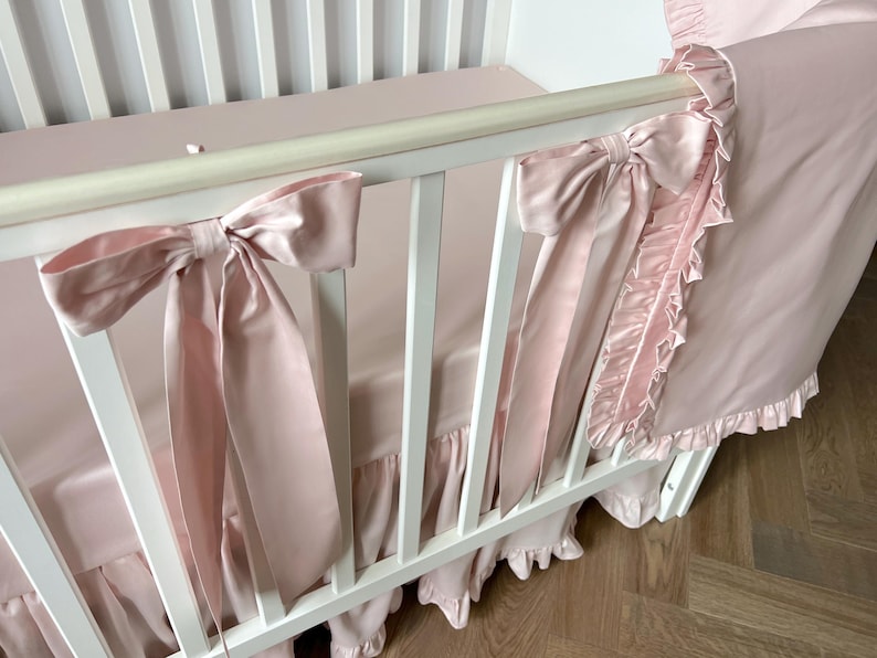 A white crib with pink ruffles and bows on the railings.
