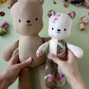 May include: Two handmade teddy bears. One is solid tan, the other is patchwork with floral patterns in pink, white, and green. Both have black button eyes and stitched smiles. The bears are held up against a light green background.