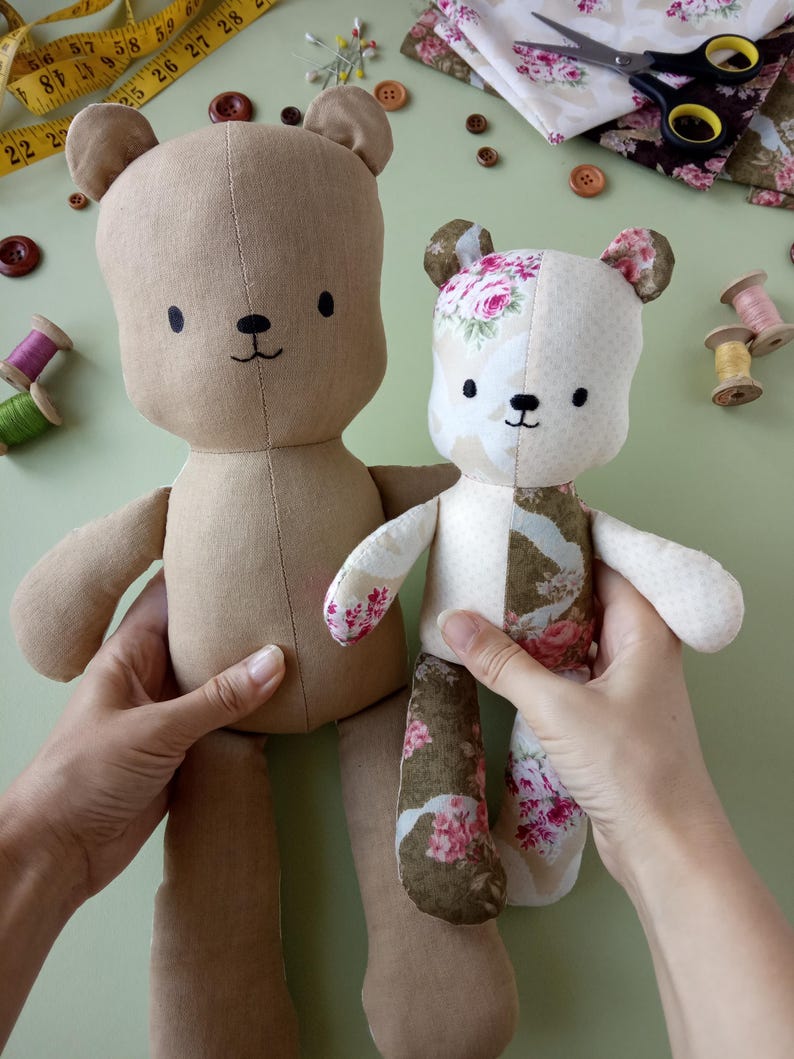 May include: Two handmade teddy bears. One is solid tan, the other is patchwork with floral patterns in pink, white, and green. Both have black button eyes and stitched smiles. The bears are held up against a light green background.