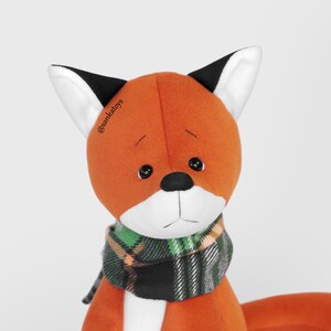 Op de afbeelding: Een pluche vos met oranje vacht, zwarte ogen en een zwarte neus. De vos draagt een groen, zwart en oranje geruite sjaal. De oren van de vos zijn wit met zwarte punten. De tekst "@sankatoys" is zichtbaar.