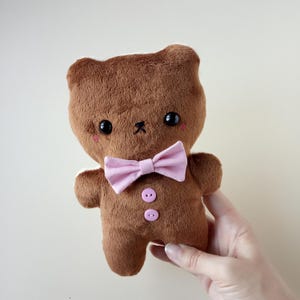 May include: A brown plush teddy bear with a pink bow tie and two pink buttons. The bear has black eyes and a small black stitched mouth. The bear is being held by a person.