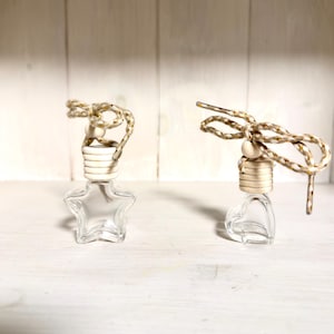Könnte beinhalten: Zwei transparente Auto-Lufterfrischer aus Glas, einer sternförmig und einer herzförmig. Jeder hat einen beigefarbenen Holzverschluss und ein dekoratives Seil mit goldenen Akzenten. Der Hintergrund ist eine weiße Holzoberfläche.