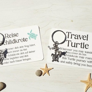 Könnte beinhalten: Zwei Schlüsselanhänger mit Schildkrötenanhängern und Karten. Auf einer Karte steht "Reise Schildkröte" und auf der anderen "Travel Turtle". Die Schlüsselanhänger haben eine silberne Oberfläche. Der Hintergrund umfasst Seesterne und Kieselsteine.