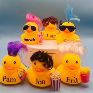 Könnte beinhalten: Eine Sammlung gelber Gummienten mit verschiedenen Frisuren, Sonnenbrillen und Accessoires. Jede Ente trägt einen Namen: Sarah, Laur, Sam, Pam, Jon und Erik. Einige Enten halten Miniaturartikel wie ein Getränk, eine Handtasche und Popcorn.