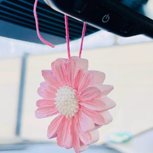 Könnte beinhalten: Ein rosa, margeritenförmiger Auto-Lufterfrischer, der an einer rosa Schnur hängt. Der Lufterfrischer hat eine weiße Mitte und rosa Blütenblätter. Der Hintergrund ist verschwommen und zeigt das Innere eines Autos.