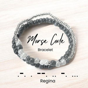 Puede incluir: Un brazalete de cuentas grises y blancas con el texto "Morse Code Bracelet" y la palabra "Regina" en código Morse debajo. El brazalete está hecho de pequeñas cuentas redondas.
