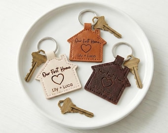May include: Three house-shaped keychains in tan, brown, and dark brown leather, each with a silver keyring and a brass key. The keychains have the text "Our First Home" and "Lily + Luca" with a heart symbol.