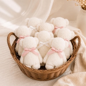 May include: A collection of small, cream-colored plush teddy bears arranged inside a round, brown woven wicker basket. Each bear features a soft, fuzzy texture and is adorned with a small, tied ribbon in either a pale pink or white color.