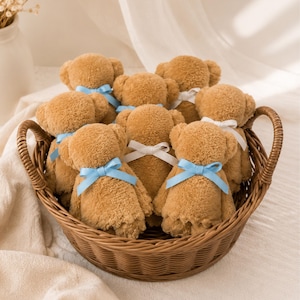 May include: A collection of small, tan, fuzzy plush teddy bears arranged inside a brown woven wicker basket. Each bear features a soft, textured surface and is adorned with a small, tied ribbon in either a light blue or white color.