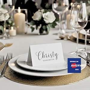 Puede incluir: Una tarjeta de sitio blanca con el nombre "Christy" en escritura elegante, colocada sobre un plato blanco. La mesa está decorada con un cargador dorado, cubiertos y copas de vino, con velas y flores en el fondo.