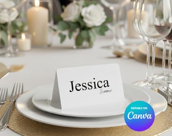 Pode incluir: Um cartão de lugar branco com o nome "Jessica" em escrita preta, colocado sobre um prato branco com um sousplat dourado. A mesa inclui talheres, taças de vinho, velas e um arranjo floral, criando um ambiente elegante.