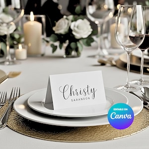 May include: A formal table setting with a white place card that reads "Christy Sarandon" in elegant script. The card sits on a white plate, atop a gold charger. Wine glasses, candles, and floral arrangements complete the elegant decor.