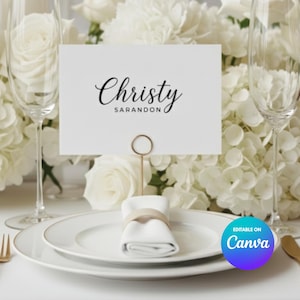 May include: A sophisticated table setting featuring white plates, a folded napkin, and a place card with the name "Christy Sarandon." The arrangement includes champagne flutes, gold cutlery, and a backdrop of white flowers, ideal for a wedding or formal dinner.