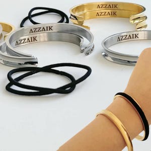 May include: Two silver and one gold bangle bracelets with the word "AZZAIk" engraved on each. The bracelets are on a white surface with a black hair tie.