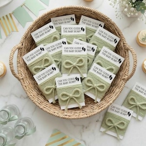 Pode incluir: Uma cesta de vime cheia de cartões de jogo para chá de bebê. Cada cartão é embrulhado com fio verde sálvia e amarrado com um laço. Os cartões dizem "CAN YOU GUESS THE BABY BUMP?" e apresentam uma pegada. A cesta está em uma superfície de mármore.