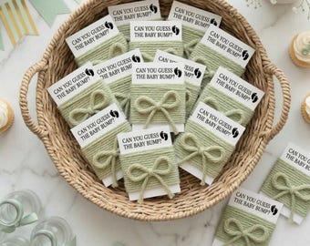 May include: A wicker basket containing baby shower game cards. Each card is wrapped in sage green yarn and tied with a bow. The cards display the text "CAN YOU GUESS THE BABY BUMP?" with a footprint graphic. The basket sits on a marble surface.