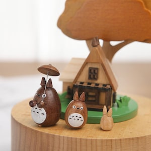 May include: Wooden music box featuring a small house, a tree, and three figurines. The figurines are of a character with an umbrella, a character with large ears, and a smaller character with ears. The music box is made of wood and has a light brown finish.