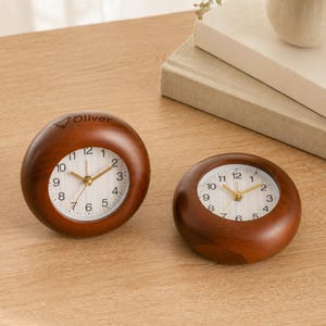 May include: Two round, dark brown wooden desk clocks with white faces, black numerals, and gold-colored hands. The larger clock on the left features the engraved name Oliver with a small heart icon above the text on the top wooden rim.