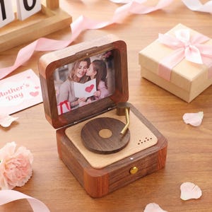 May include: A wooden music box resembling a vintage record player, with a photograph of a mother and child inside. A small gift box tied with a pink ribbon and a "Mother's Day" card are also present.