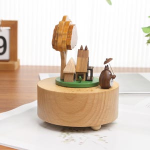 May include: Wooden music box with a small house, tree, and a brown character holding an umbrella. The music box is made of light brown wood and has a green base.