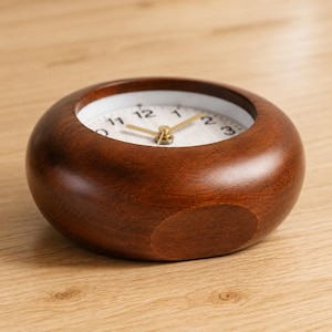 May include: A round, dark brown wooden desk clock with a smooth, polished finish. The clock face is white with black numerals from 1 to 12 and features thin, gold-colored hour, minute, and second hands centered within the circular wooden frame.