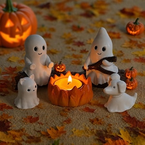 Peut inclure: Un ensemble de figurines fantômes en céramique blanche, un fantôme chat blanc et un fantôme chien blanc disposés autour d'un bougeoir central en céramique orange en forme de feu de camp, avec de petites décorations citrouilles orange.