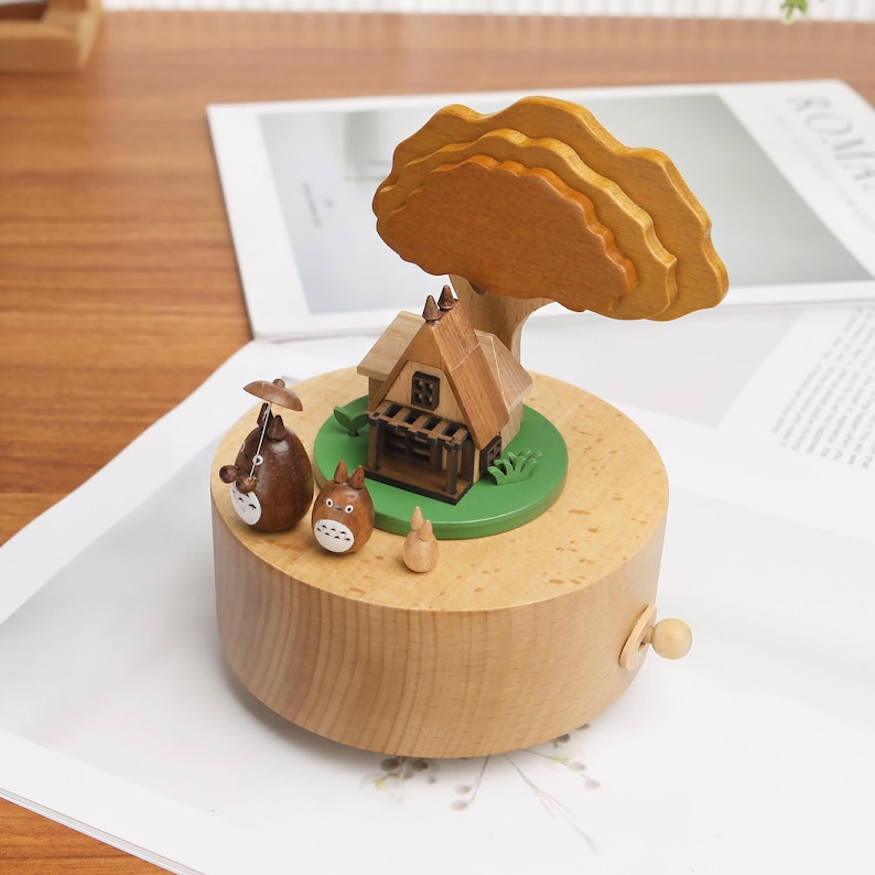 May include: A wooden music box with a small house, a tree, and two cartoon characters. The music box is brown and has a green base. The house is brown and has a white door and windows. The tree is brown and has green leaves. The characters are brown and have white eyes and noses.