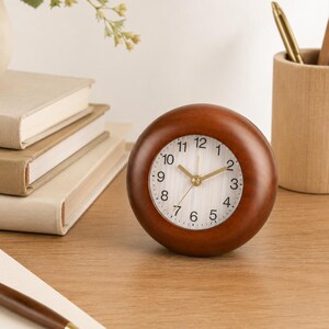 May include: A round analog desk clock with a polished dark brown wooden frame. The clock face is white with vertical pinstripes, featuring black Arabic numerals from 1 to 12 and gold-colored hour, minute, and second hands centered on the dial.