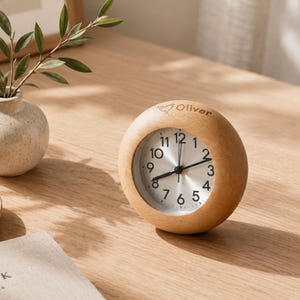 May include: A round wooden alarm clock with a light brown finish and a silver face featuring black numerals. The top of the wooden frame is engraved with a small heart icon and the name Oliver. The clock has black hour, minute, and second hands.