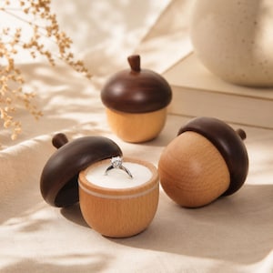 May include: Three acorn-shaped wooden ring boxes with dark brown caps and light tan bases. One box is open, revealing a white interior cushion holding a silver ring with a clear stone. The other two boxes are closed, showing smooth wood grain.