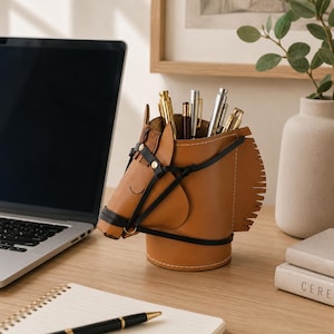 Può includere: Un portapenne in pelle color cuoio a forma di testa di cavallo, dotato di una briglia in pelle nera con finiture metalliche, criniera sfrangiata e base cilindrica. Il design presenta dettagli cuciti e contiene diverse penne metalliche.