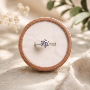 May include: A silver-toned ring featuring a single round-cut clear gemstone held in a six-prong setting. The ring is centered within a circular wooden display box that has a soft, off-white velvet interior lining for holding jewelry securely.