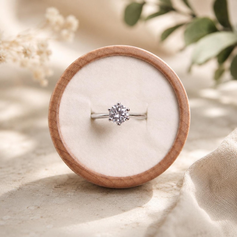 May include: A silver-toned ring featuring a single round-cut clear gemstone held in a six-prong setting. The ring is centered within a circular wooden display box that has a soft, off-white velvet interior lining for holding jewelry securely.