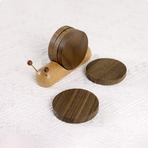 May include: A wooden snail-shaped coaster holder with four dark brown coasters. The snail's body is light-colored wood, and the coasters are round. The snail has two antennae with small brown balls at the ends.