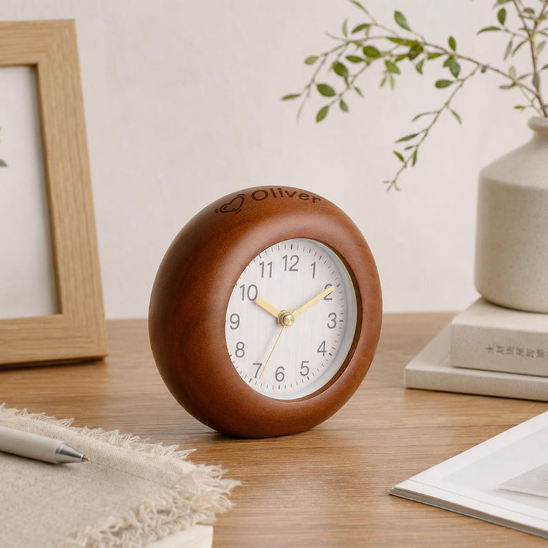 May include: A round wooden desk clock with a smooth, dark brown finish and a white face featuring black numerals. The top of the wooden frame is engraved with the name Oliver and a small heart icon. The clock has thin, gold-colored hands.
