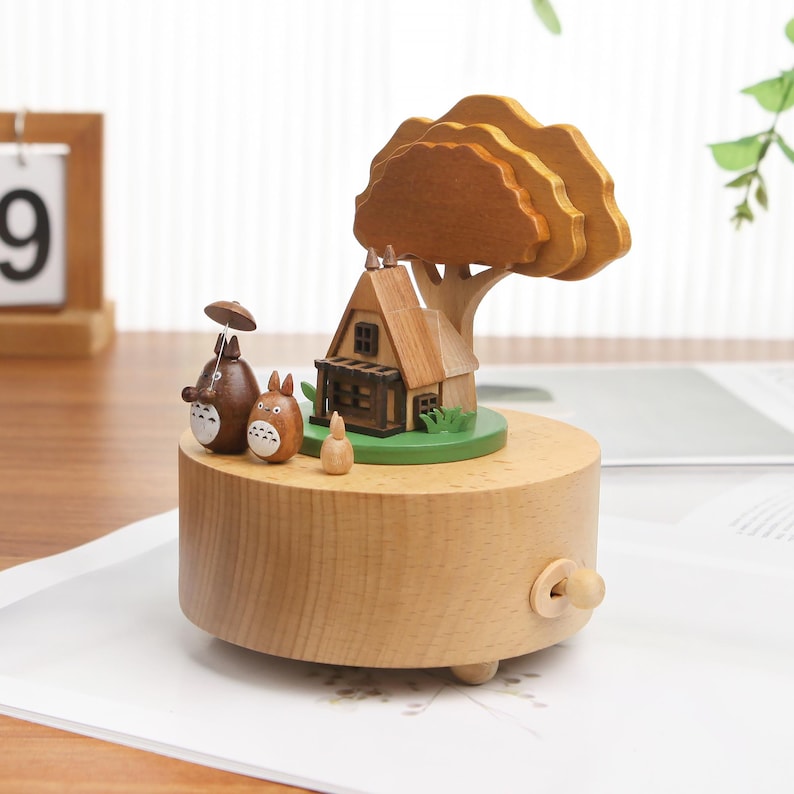 May include: Wooden music box with a small house, tree, and two cartoon characters. The music box is brown and has a crank on the side.