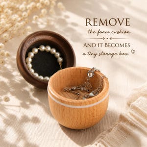 May include: A round wooden jewelry box with a light wood base and a dark wood lid. The lid contains a white pearl ring on a black foam cushion. The base holds a silver chain necklace. Text reads: REMOVE the foam cushion AND IT BECOMES a tiny storage box.
