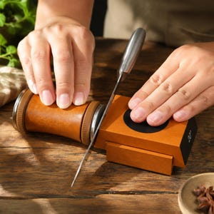May include: A wooden knife sharpening tool with a cylindrical handle and a rectangular base featuring a black circular inlay and a 20-degree angle indicator. The device is constructed from light brown wood with metallic accents and a silver blade.