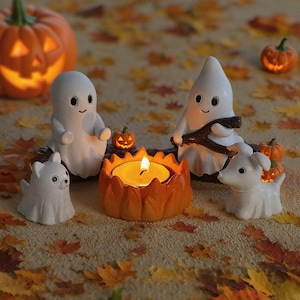 May include: A Halloween-themed display featuring a lit pumpkin-shaped candle holder, white ghost figurines, and small pumpkin decorations. The scene is set on a surface covered with autumn leaves, creating a festive atmosphere.
