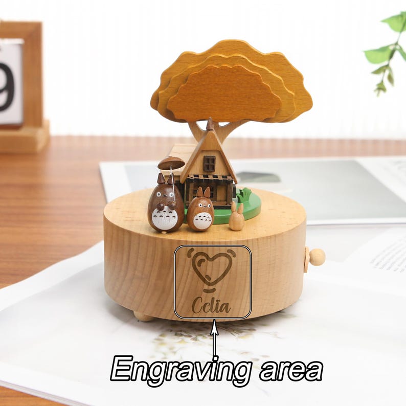 May include: Wooden music box with a small house, tree, and two cartoon characters. The music box has a heart and the name "Celia" engraved on the front.
