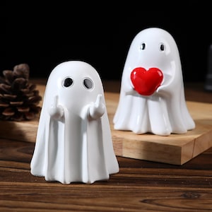 Puede incluir: Dos figuras de fantasmas de cerámica blanca. Un fantasma hace la peineta y el otro sostiene un corazón rojo. Las figuras están sobre una superficie de madera.