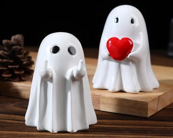 Puede incluir: Dos figuras de fantasmas de cerámica blanca. Un fantasma hace la peineta y el otro sostiene un corazón rojo. Las figuras están sobre una superficie de madera.