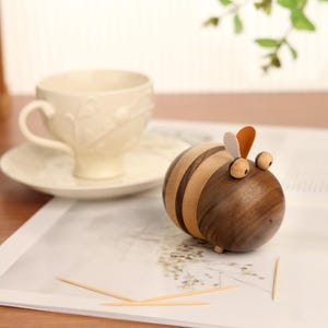 May include: A wooden bee-shaped toothpick holder with a striped body, brown wings, and black eyes. The holder is sitting on a white surface with toothpicks beside it.