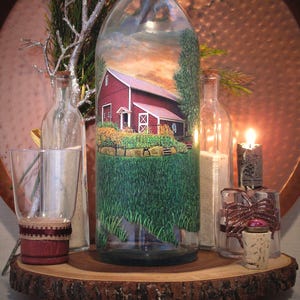 May include: A decorative glass bottle with a painted rural scene of a red barn, green grass, and a sunset sky. The bottle is surrounded by other glass bottles, a candle, and a glass with a decorative band, all arranged on a wooden slice.
