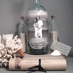 May include: A decorative unity sand ceremony set. The set includes a large glass bottle with a photo of a couple on the beach, two smaller bottles, sand, a scroll, and a seashell. The large bottle has a cork stopper.
