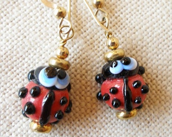 May include: Gold earrings with red and black ladybug charms. The ladybugs have black spots, white eyes, and blue pupils.