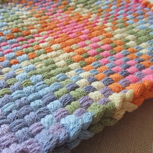 May include: A woven square mat with a colorful pattern. The mat is made of different colored yarn, including blue, green, purple, orange, pink, and cream.